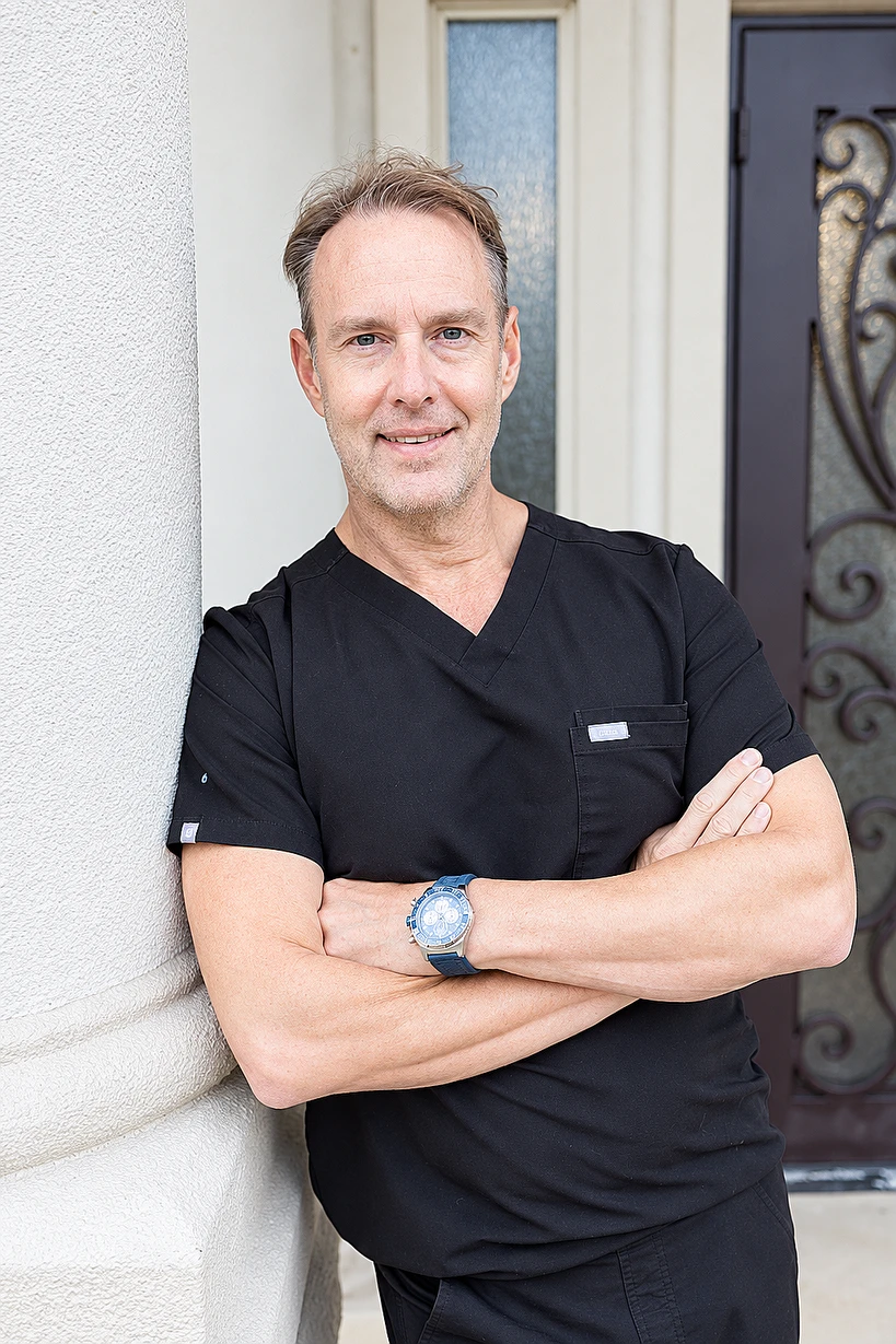 Dr. Tom Draper in black scrubs, arms folded, leaning on a stone column