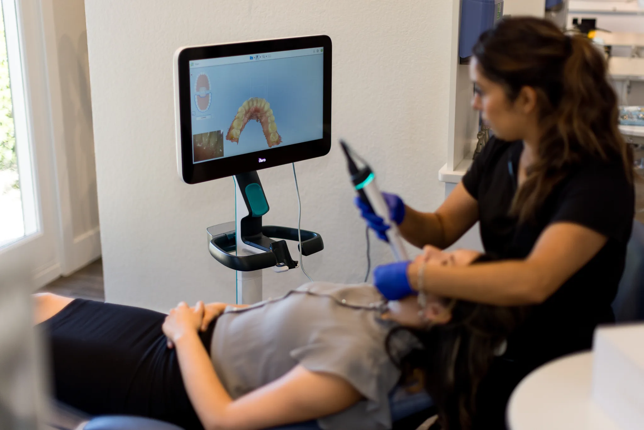 A dental assistant passing an intraoral scanner over a patient's teeth while the 3D model builds on the chairside monitor