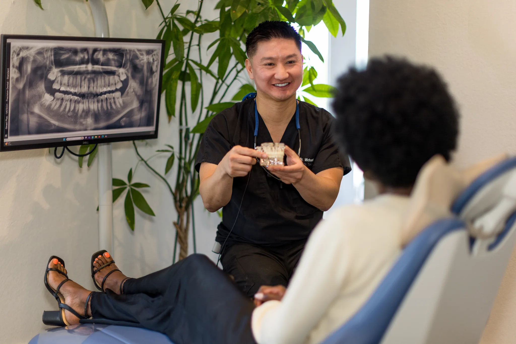 Dr. Nguyen holding an implant model while talking with a seated patient, a panoramic X-ray on the wall behind