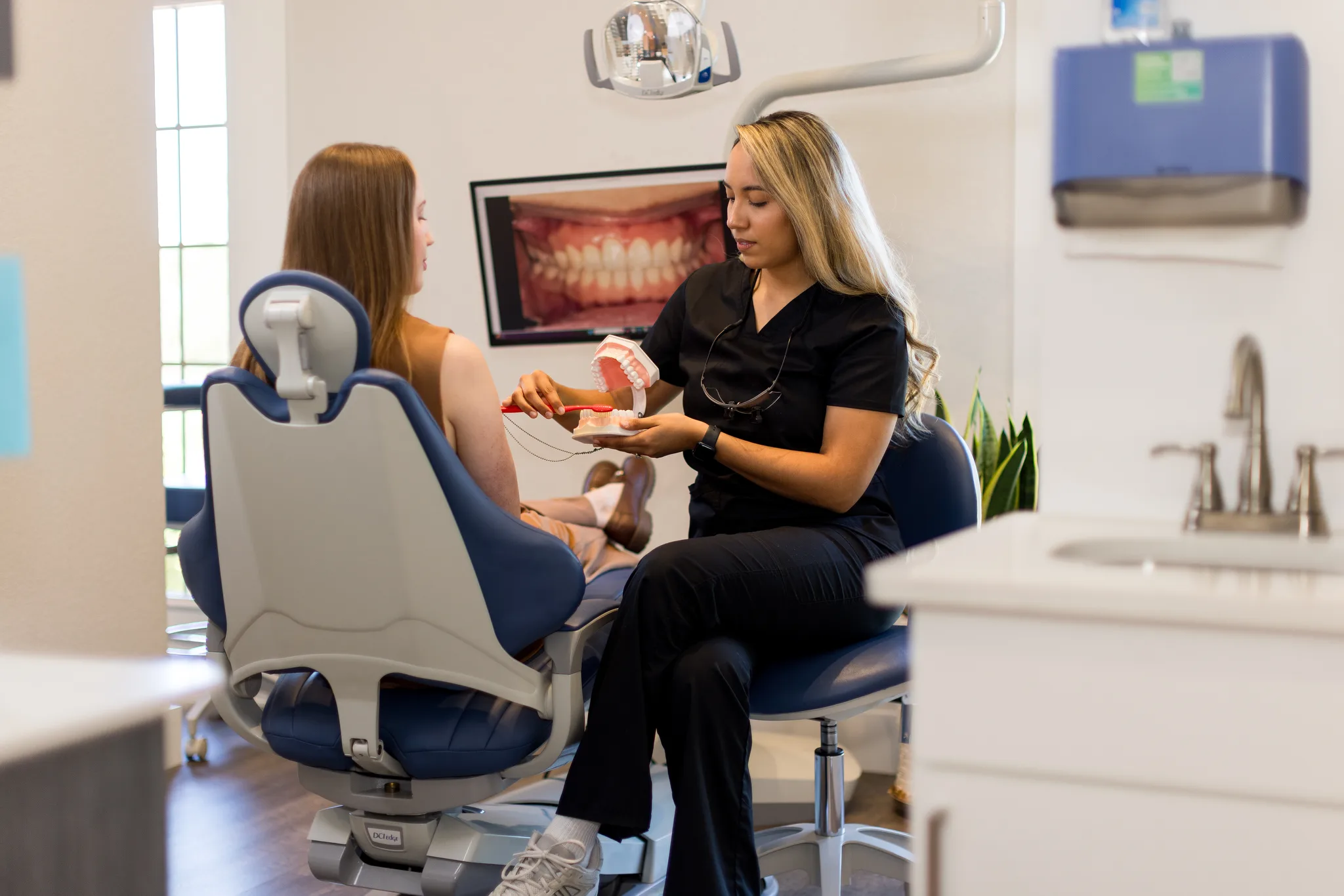 A hygienist holding a model beside a patient while intraoral photographs show on screen