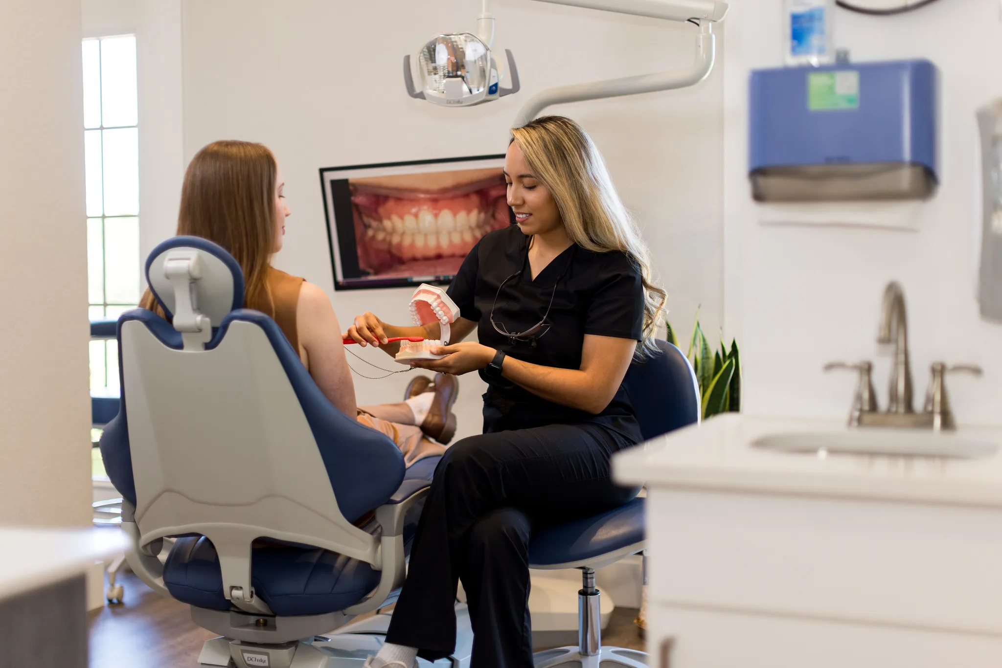A hygienist using a model to explain treatment, with intraoral photographs on the screen behind